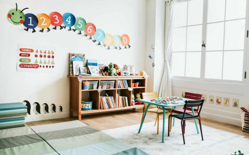 jasna sala przedszkolna, na ścianie gąsiennica z cyferek, półka z książkami i stolik dla dzieci. Jak wybrać przedszkole