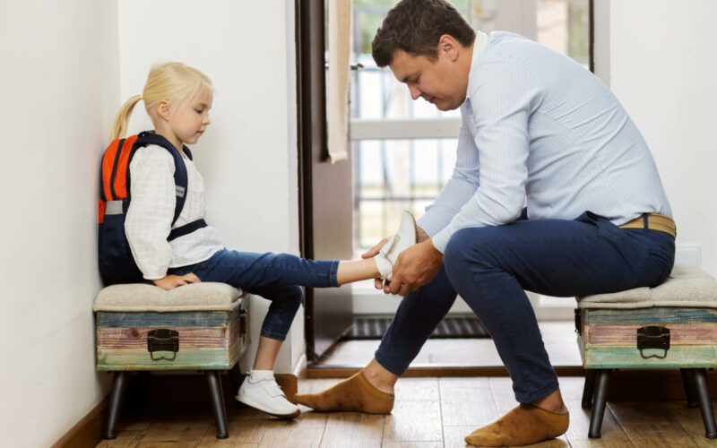 Samodzielność w przedszkolu – tata zakłada buty małej dziewczynce z plecakiem