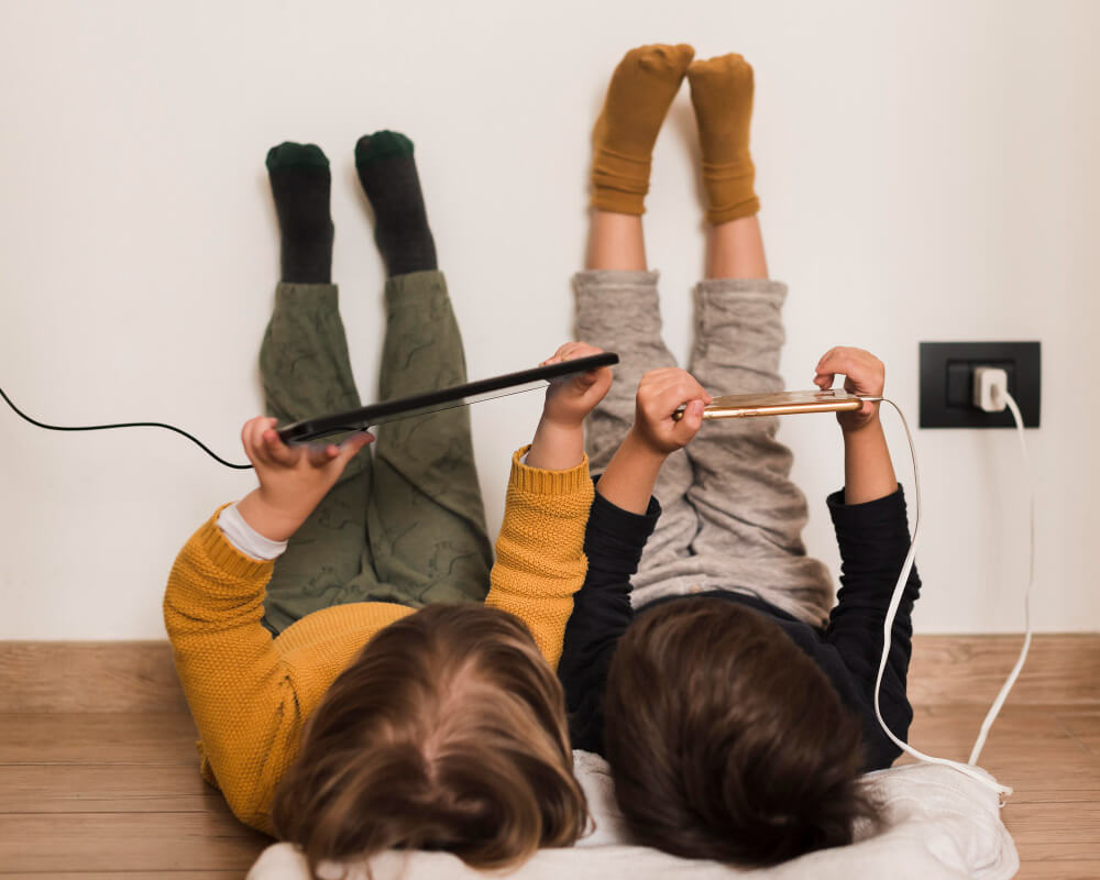 Dzieci leżą na podłodze trzymając podłączone do ładowarki smartfony. Ile czasu dziecko może spędzać przed ekranem?